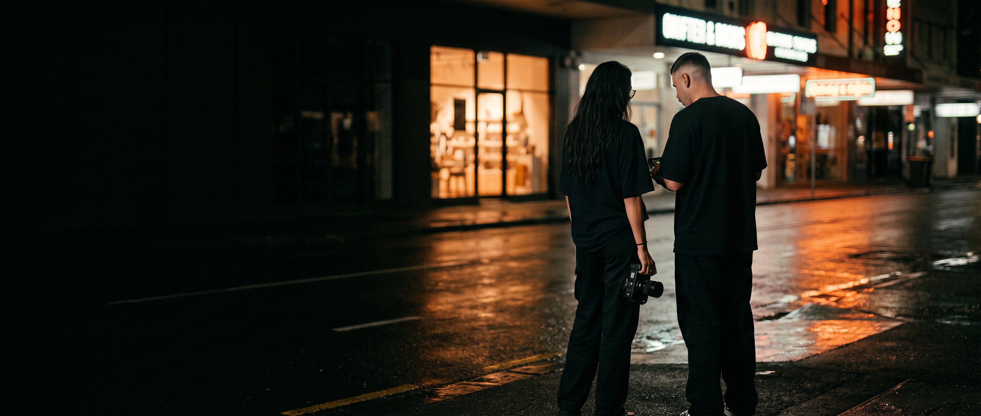 A woman and a man on a dark city street at night, backs to camera, one holding a camera.