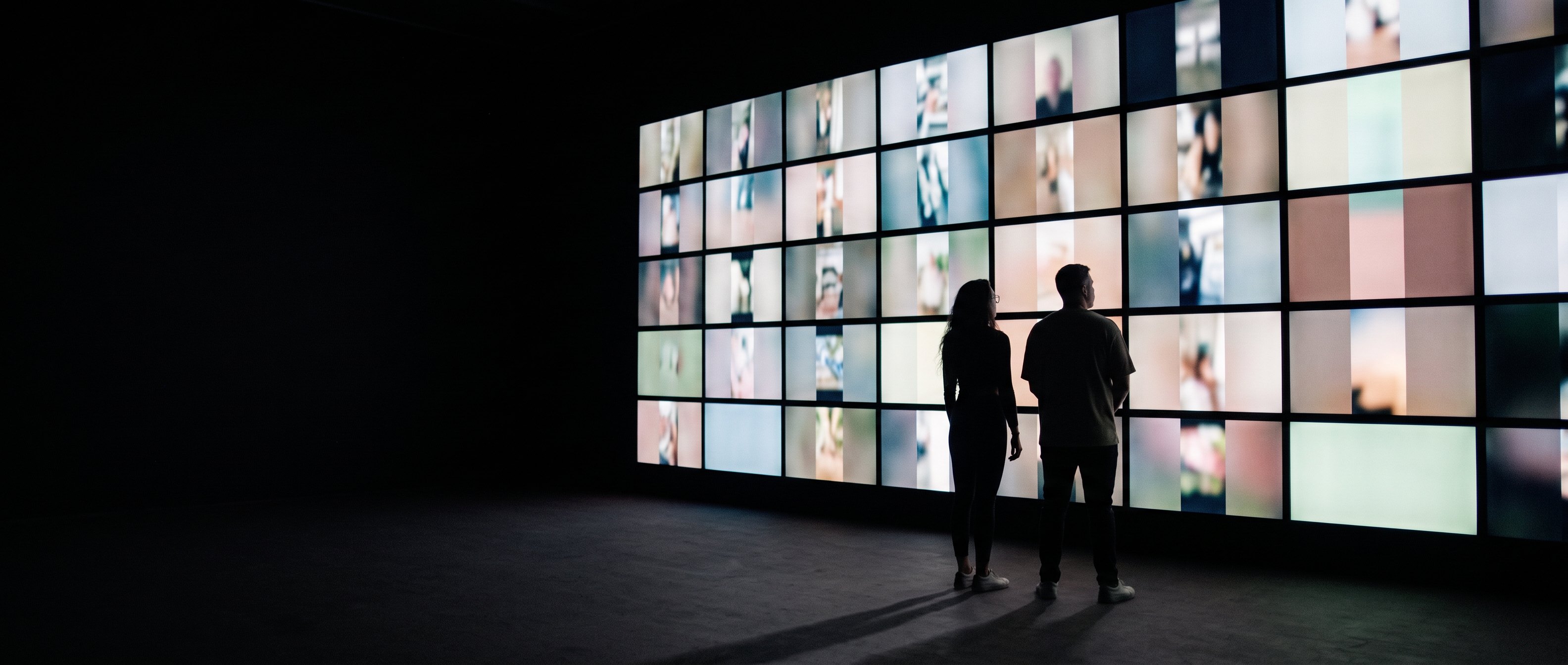 Two people seen from behind in a dark room, facing a large wall of glowing video screens.
