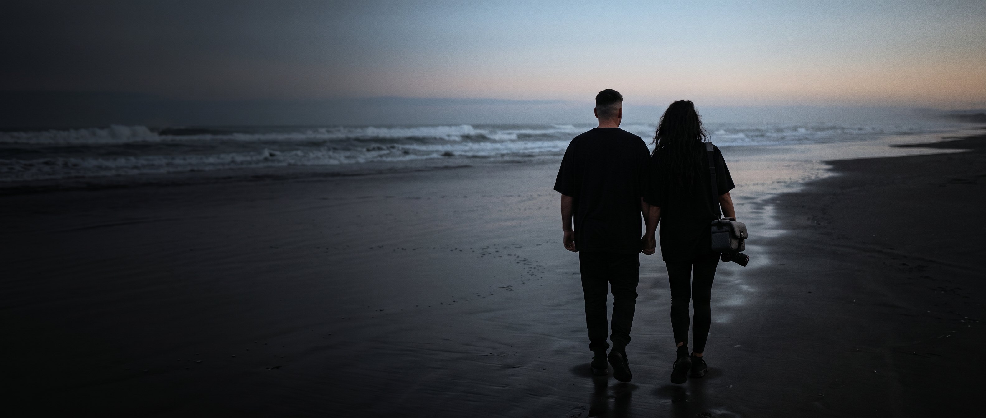 A man and a woman walking along a beach at dusk, backs to camera, one carrying a camera.